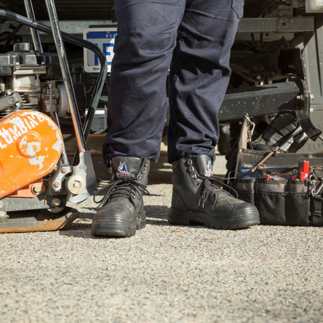 A close-up of a person's legs from the knees down, wearing black leather work boots with white and blue 'Crown' branding, standing on asphalt next to a piece of heavy machinery and a tool belt.